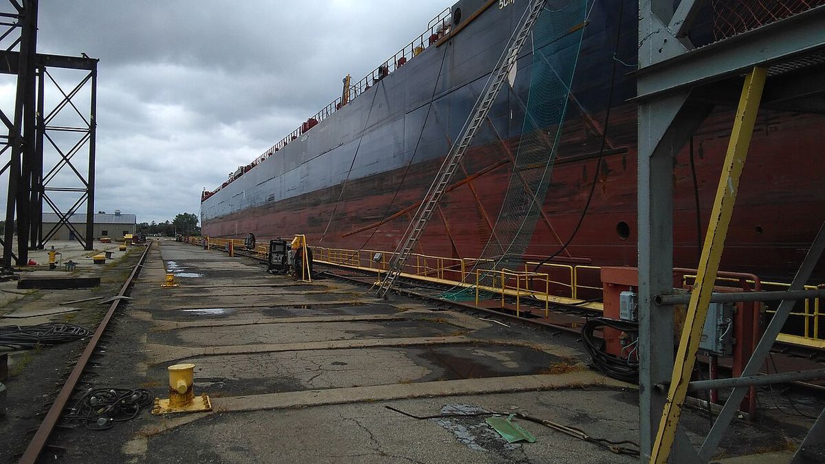 A Great Lakes freighter in a dry dock at a shipyard, with access ladders and safety netting along the hull