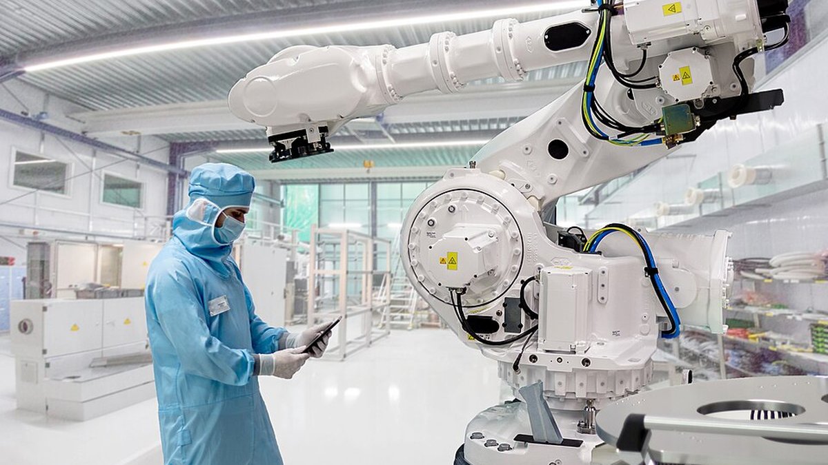 Technician in a cleanroom suit checking a tablet beside a large white industrial robot arm